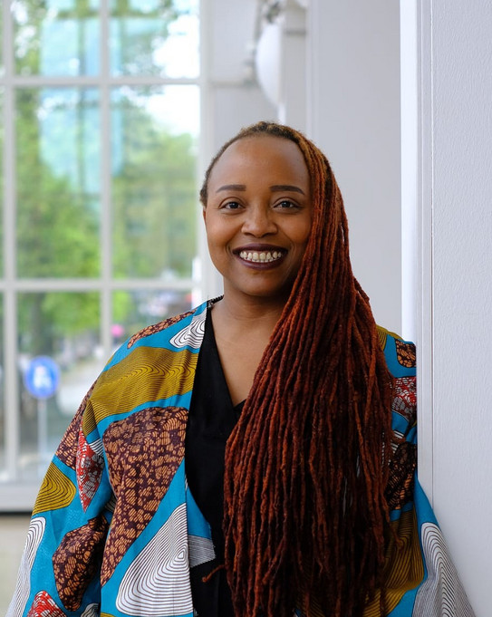 Foto von Person Dr. Rahab Njeri mit langen Dreadlocks und bunter Jacke lehnt an einer weißen Wand vor einem großen Fenster