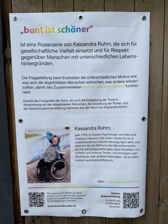 Ein Plakat mit weißem Hintergrund und schwarzem Text, oben mittig steht der farbige Schriftzug 'bunt ist schöner'. Darunter folgt ein erklärender Text zur Postkartenserie von Kassandra Ruhm, die sich für gesellschaftliche Vielfalt und Respekt gegenüber Menschen mit unterschiedlichen Lebenshintergründen einsetzt. Im unteren Bereich befindet sich ein Foto einer Person im Rollstuhl, die draußen auf einem Weg sitzt und mit einer Hand winkt. Rechts neben dem Foto steht ein kurzer Lebenslauf von Kassandra Ruhm. Am unteren Rand sind zwei QR-Codes und eine Webadresse abgebildet.
