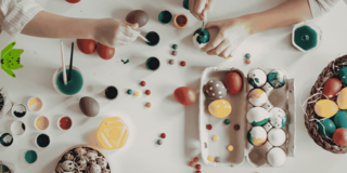 Zwei Hände bemalen bunte Ostereier auf einem Tisch mit Farbtöpfen und bemalten Eiern in einem Eierkarton und einem Korb